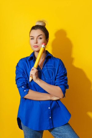 Photo for Cheerful young woman wearing blue shirt holding giant pencil on yellow background - Royalty Free Image