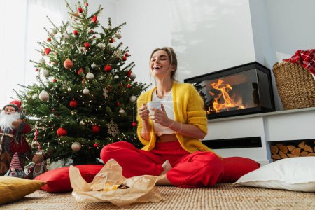 Photo for Happy woman sits beside a glowing fireplace in a cozy living room, adorned with a Christmas tree and festive decorations, enjoying a cup of hot drink. - Royalty Free Image