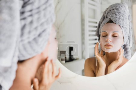Téléchargez les photos : Belle femme dans la salle de bain avec masque de feuille appliquée sur son visage regardant dans le miroir - en image libre de droit