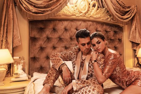 Téléchargez les photos : Le jeune couple riche est vêtu de tenues dorées rayonnantes ornées de broderies à paillettes, situées dans une suite luxueuse. - en image libre de droit