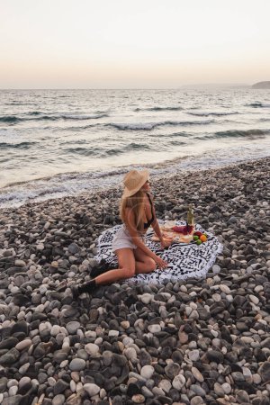 Téléchargez les photos : Femme élégante et insouciante portant un chapeau de paille fait un pique-nique serein sur une plage de galets pendant un coucher de soleil. - en image libre de droit