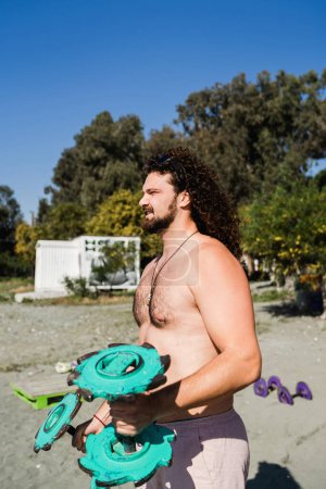 Foto de Hombre rizado haciendo ejercicio con pesas de bricolaje en el gimnasio de playa al aire libre - Imagen libre de derechos