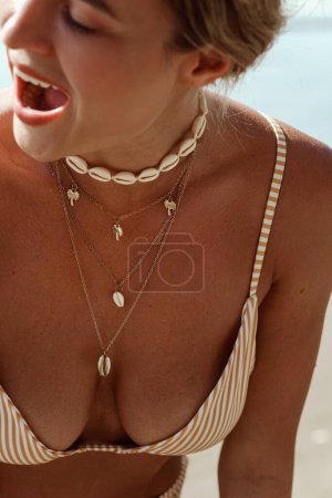 Photo for Cheerful woman in bikini wearing golden necklace with palm trees and seashells on a sunlit beach. - Royalty Free Image