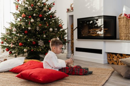 Téléchargez les photos : Mignon petit garçon joue à côté d'une cheminée dans un salon confortable orné d'un arbre de Noël et de décorations festives. - en image libre de droit
