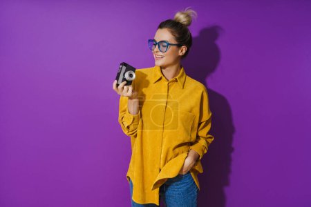 Photo for Portrait of young cheerful girl wearing eyeglasses holding vintage film camera on purple background - Royalty Free Image