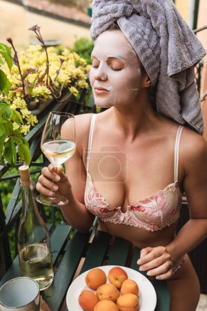 Foto de Retrato de una joven y hermosa mujer con lencería con máscara de sábana facial aplicada en su cara sentada en un balcón durante su desayuno - Imagen libre de derechos