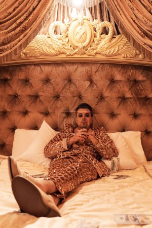 Téléchargez les photos : Homme riche et excentrique vêtu d'un costume doré chatoyant avec beaucoup de billets en dollars dans une suite royale et luxueuse. - en image libre de droit