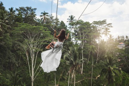 Foto de Mujer joven con vestido blanco balanceándose en columpios de cuerda con hermosa vista sobre terrazas de arroz y palmeras en la isla de Bali - Imagen libre de derechos