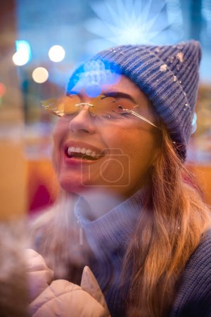 Photo for Cheerful and stylish woman, dressed in warm clothes and wearing sunglasses shaped like fire flames, is having fun in a snowy winter amusement park. - Royalty Free Image