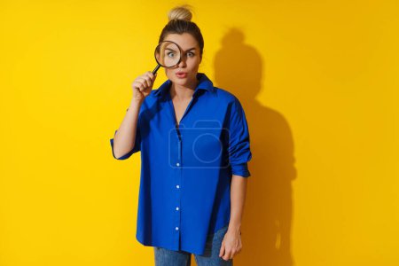 Foto de Mujer curiosa está mirando a través de lupa con gran interés, examinando algo de cerca en el fondo amarillo. - Imagen libre de derechos