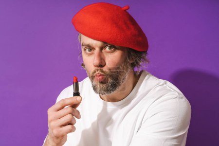 Photo for Funny middle aged man wearing red beret is holding lipstick in his hand against purple background. - Royalty Free Image