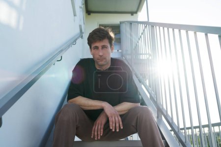 Téléchargez les photos : Portrait d'un jeune homme adulte sérieux portant des vêtements de sport assis dans les escaliers près de sa maison - en image libre de droit