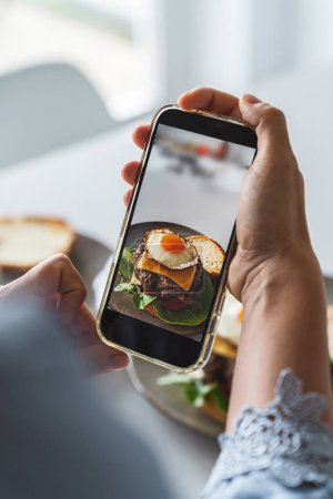 Foto de Manos femeninas usando un smartphone para fotografiar una deliciosa hamburguesa casera con queso y un huevo frito encima. - Imagen libre de derechos
