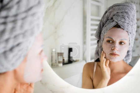 Foto de Hermosa mujer en el baño con máscara de hoja aplicada en su cara mirando en el espejo - Imagen libre de derechos