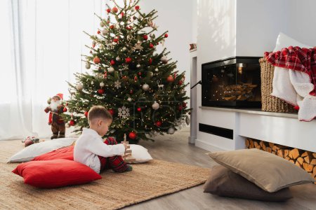 Téléchargez les photos : Mignon petit garçon joue à côté d'une cheminée dans un salon confortable orné d'un arbre de Noël et de décorations festives. - en image libre de droit