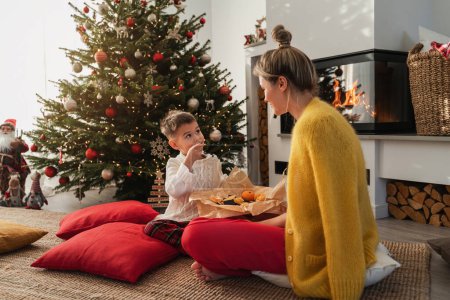 Téléchargez les photos : Jeune femme et son petit fils assis à côté d'une cheminée dans un salon confortable, orné d'un arbre de Noël et de décorations festives, profitant de quelques bonbons. - en image libre de droit