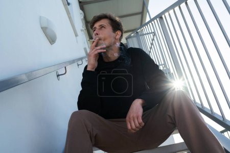 Foto de Hombre joven adulto fumando cigarrillo RYO o marihuana en la escalera junto a su casa - Imagen libre de derechos