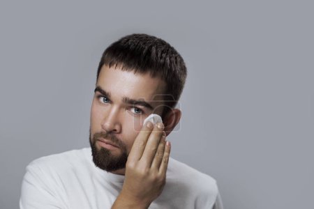 Téléchargez les photos : Jeune homme beau nettoie son visage avec un tampon de coton sur fond gris - en image libre de droit