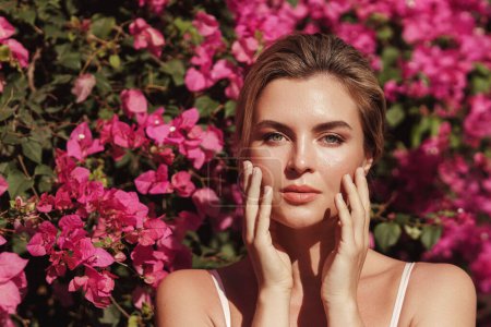 Foto de Hermosa mujer rubia con piel lisa y maquillaje natural, mientras posa sobre un telón de fondo de un arbusto con delicadas flores rosadas. - Imagen libre de derechos