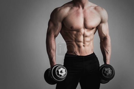 Photo for Young and muscular fitness man with dumbbells, showcases his muscular chest, abs and arms against gray background. - Royalty Free Image