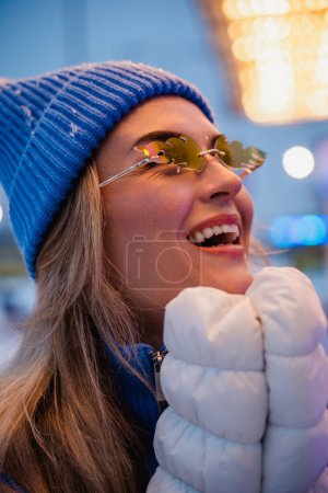 Foto de Mujer alegre y elegante, vestida con ropa de abrigo y con gafas de sol en forma de llamas de fuego, se divierte en un parque de atracciones de invierno nevado. - Imagen libre de derechos