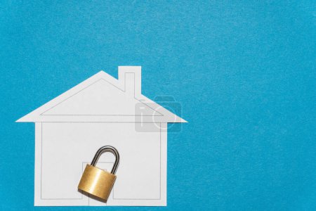 Téléchargez les photos : Serrure métallique jaune devant une forme découpée de maison en papier sur fond bleu - en image libre de droit