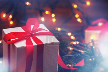 Foto de Cajas de regalo envueltas con cintas rojas vibrantes, enclavadas entre luces centelleantes de Navidad y ramitas de abeto fresco. - Imagen libre de derechos