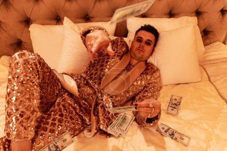 Foto de Hombre rico y excéntrico vestido con un brillante traje dorado con muchos billetes de dólar en una suite real y lujosa. - Imagen libre de derechos