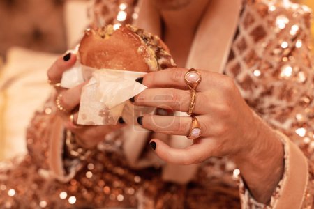 Téléchargez les photos : Homme riche dans un costume doré chatoyant portant des anneaux d'or et mangeant un hamburger. - en image libre de droit