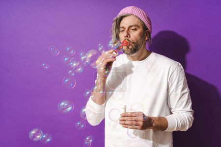 Téléchargez les photos : Insouciant homme d'âge moyen soufflant des bulles de savon sur fond violet. - en image libre de droit