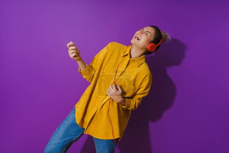 Téléchargez les photos : Portrait d'une jeune fille joyeuse portant une chemise jaune écoutant de la musique à l'aide d'écouteurs rouges et jouant de la guitare à air sur fond violet - en image libre de droit