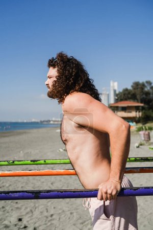 Photo for Curly and bearded man working out on parallel bars during his beach training - Royalty Free Image