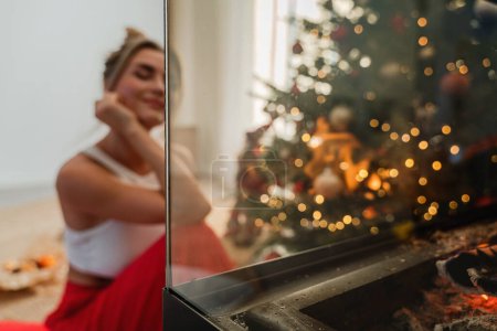 Photo for Young woman sits beside a glowing fireplace in a cozy living room adorned with a Christmas tree and festive decorations. - Royalty Free Image