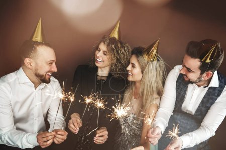 Photo for Group of happy people wearing party hats holding burning sparkles during holiday or event celebration - Royalty Free Image