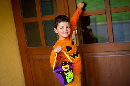 Téléchargez les photos : Un garçon de sept ans habillé en citrouille et portant un sac de bonbons frappe à la porte et sourit. - en image libre de droit