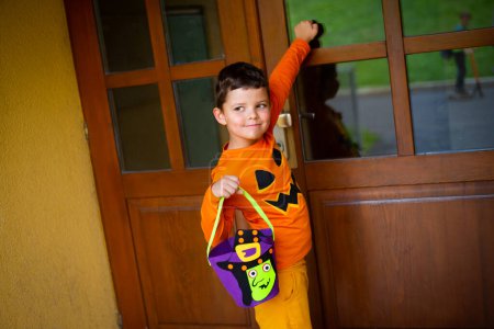 Téléchargez les photos : Un garçon de sept ans habillé en citrouille et portant un sac de bonbons frappe à la porte et sourit. - en image libre de droit