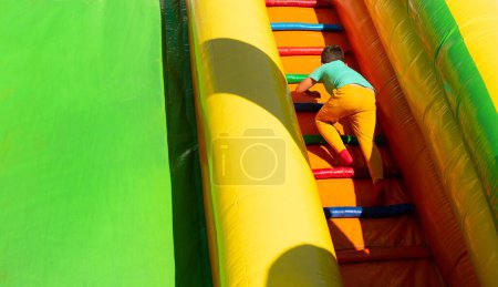 Photo for Little boy child in a turquoise T-shirt and yellow mustard pants climbs the stairs of a multi-colored slide in the park. - Royalty Free Image