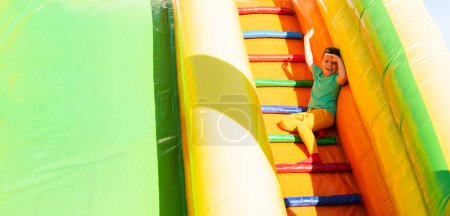 Foto de Un niño pequeño con ropa de colores brillantes se sienta, sonríe y agita las escaleras de un colorido tobogán en el parque en un día de verano muy soleado. Cubre sus ojos con su mano del sol - Imagen libre de derechos