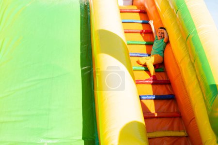 Photo for A little boy in bright colored clothes sit, smile and wave on the stairs of a colorful slide in the park on a very sunny summer day. He cover his eyes with his hand from the sun - Royalty Free Image