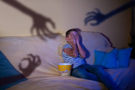Foto de Un niño pequeño está viendo una película de terror, sentado en el sofá con palomitas de maíz, cubriendo sus ojos con sus manos, y en la pared las sombras de las manos de miedo están llegando a él - Imagen libre de derechos