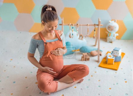 Pregnant woman smiling at belly in nursery playrooom with Baby activity gym play, toys and playmat. Pregnancy concept and home nusery planning and decoration concept photo with happy expecting woman.