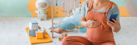 Pregnant woman belly in nursery playrooom using phone app. Pregnancy concept and home nusery planning and decoration concept. Unrecognizable women in panoramic banner.