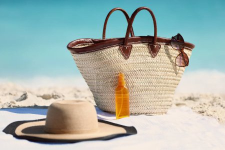Womens beach accessories on sand for summer vacation concept. Straw tote bag, sun hat and sunscreen lotion or suntan tanning oil spray bottle with blue ocean background for travel holidays.