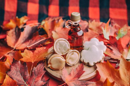 Maple syrup butter and taffy cones with leaf shape cookies on traditional Quebec plaid table with red autumn leaves. Small gift bottle. Traditional food.