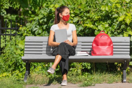 University student wearing mask sitting on bench going back to school on campus holding laptop. Asian woman with backpack, books, looking away waiting for bus. Coronavirus pandemic.