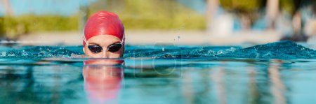 Swimming athlete man creative portrait wearing swim goggles and swim cap in swimming pool while doing breast stroke. Male swimmer swimming breaststroke in pool outside. Panoramic banner.