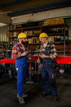 Arbeiter in einer mechanischen Werkstatt sprechen miteinander, während sie versuchen, eine Lösung zu finden, um eine Landmaschine zu starten. Beide tragen Arbeitskleidung und langärmeliges Hemd sowie Helme.