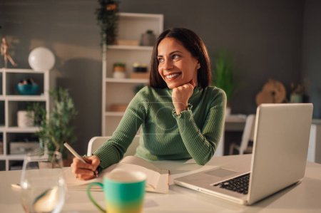 Une femme autodidacte joyeuse est assise au bureau à la maison et suit des cours en ligne et des webinaires tout en écrivant dans un manuel. Une femme utilise des plateformes d'apprentissage en ligne pour l'éducation et l'apprentissage.