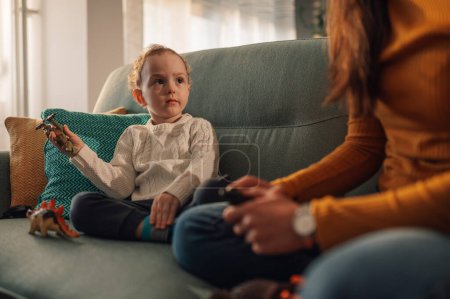 Ein Sohn spielt mit seiner Mutter mit Dinosaurierspielzeug, während er zu Hause auf dem Sofa sitzt. Ein nachdenklicher Junge spielt mit seinen Eltern in einer gemütlichen Wohnung und verbringt viel Zeit miteinander.