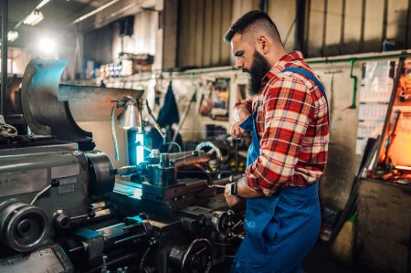 Ein junger Maschinist mit Bart steht neben einer Drehmaschine und bastelt. Er repariert die Maschine in einer Fabrik. Unscharfer Hintergrund. Er trägt Overalls und ein rotes kariertes Hemd.